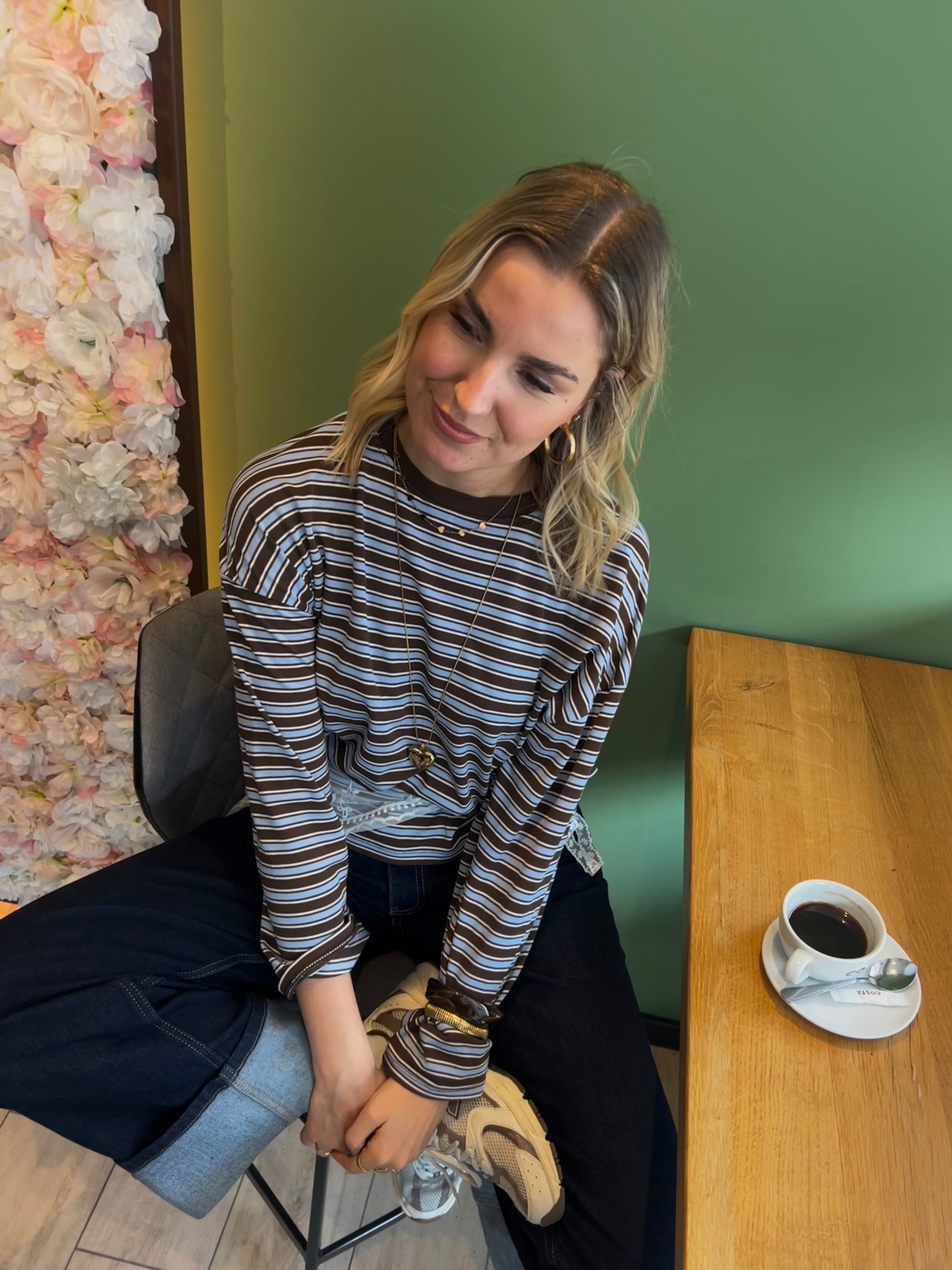 Entspannt sitzt eine Frau im gestreiften Shirt in einem gemütlichen Café. Beworben wird die neue Frühjahrskollektion 2026 von Dutch Atelier. Niederländische Mode.
