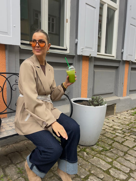 Yarah Blazer aus dem Onlineshop von Dutch Atelier sitzt mit einem Smoothie in der Sonne auf einer Bank. 